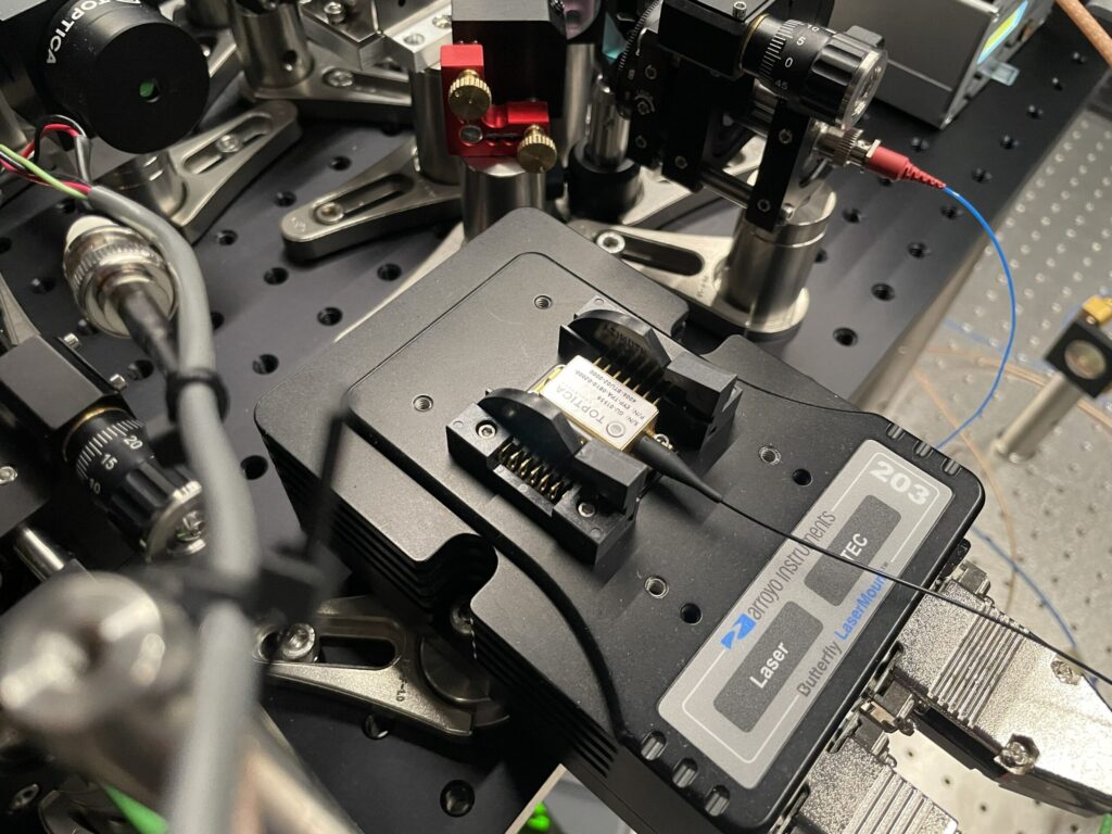 Close-up of scientific lab equipment featuring a laser diode module connected to various mounts and optical components on an optical table.